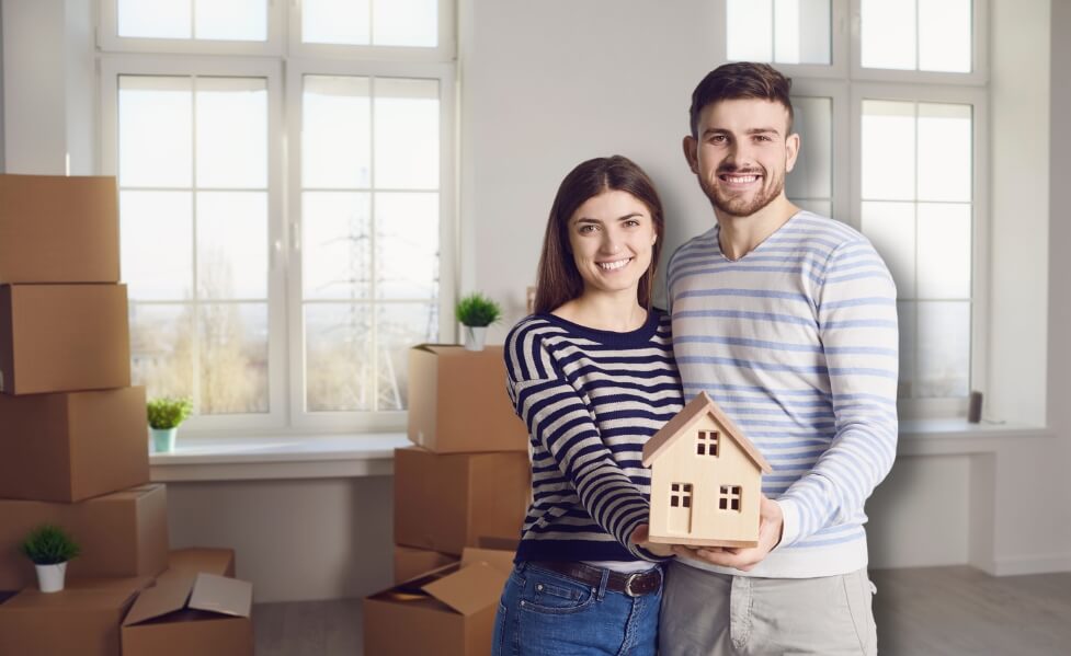Ein junges Paar steht fröhlich im neuen Zuhause, im Hintergrund stehen noch geschlossene Packkartons